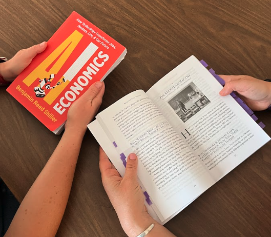 Two people reading an open copy of AI Economics at a wooden table, with the book cover visible beside them
