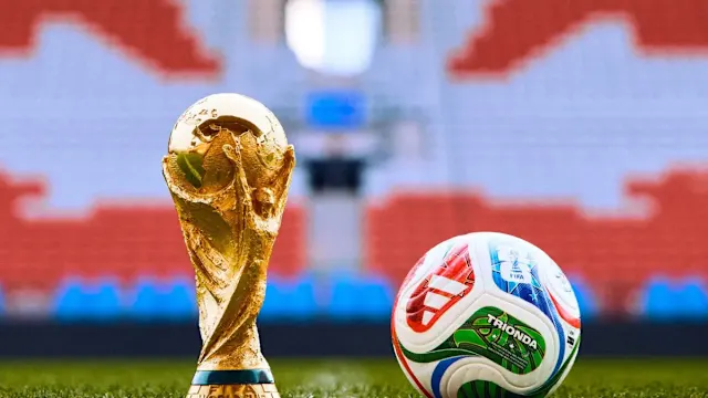 The FIFA World Cup trophy stands beside an official soccer ball on a grass pitch with a stadium in the background.