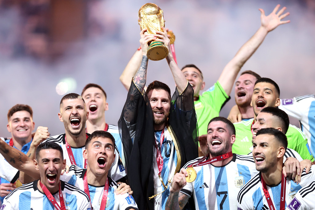 Argentina's national soccer team celebrates winning the 2022 FIFA World Cup, with Lionel Messi lifting the trophy above his head while teammates cheer around him wearing gold medals.