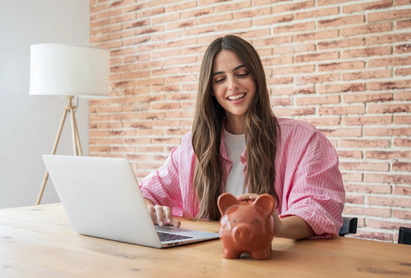 A smiling young woman in a pink striped shirt sits at a wooden table in front of a brick wall, typing on a laptop with one hand while resting her other hand on top of a terracotta piggy bank.