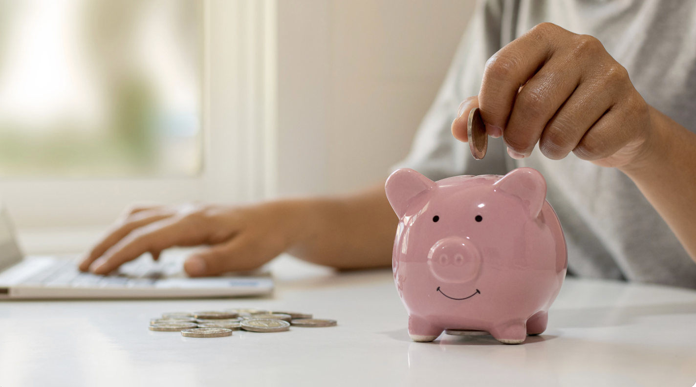 A person drops a coin into a smiling pink piggy bank with one hand while typing on a laptop with the other, and a small pile of coins sits beside them on a white desk near a bright window.