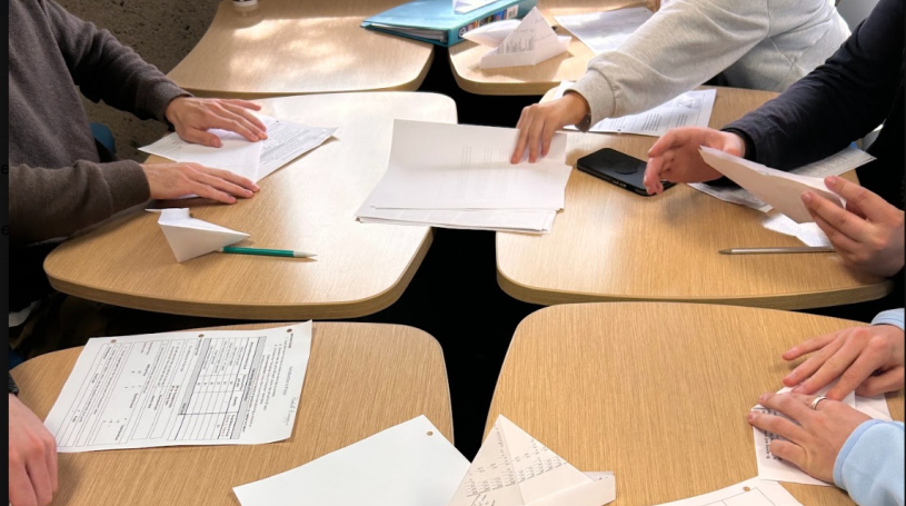 Students seated at grouped desks with folded paper products, folding instruction handouts, and cost worksheets spread in front of them.