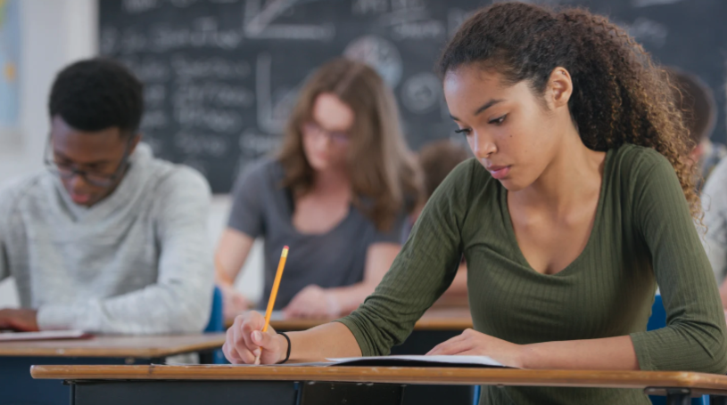 Students working independently at their desks, writing at an assessment in a classroom with a chalkboard in the background.