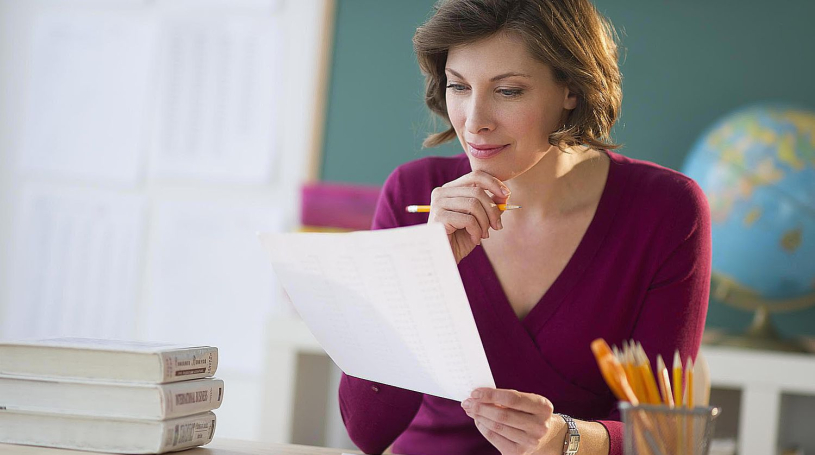 Teacher seated at a desk holding a printed page of data, pencil in hand, reviewing it closely.