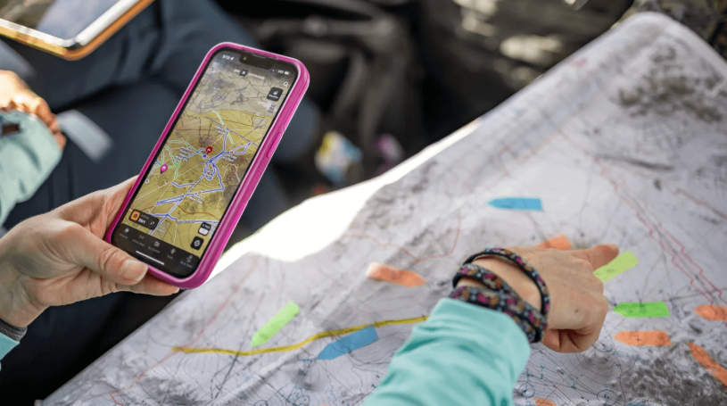 A person points at a paper topographic map marked with colored highlighter while holding a phone that shows the same terrain in a digital mapping app. Image: Garth Milan