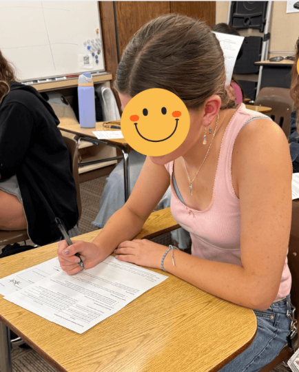 High school students completing a written in-class economics assessment at individual desks, with student faces obscured for privacy.