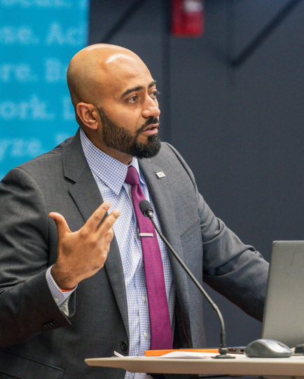 Abdullah Al Bahrani, creator of Decode Econ, speaking at a podium, gesturing with one hand mid-sentence, with a laptop and microphone in front of him.