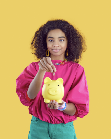 A smiling teenage girl with curly hair, wearing a bright pink top and green pants, drops a coin into a yellow piggy bank that she holds in her open palm against a solid yellow background.