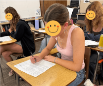 High school students completing a written in-class economics assessment at individual desks, with student faces obscured for privacy.