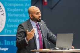 Abdullah Al Bahrani, creator of Decode Econ, speaking at a podium, gesturing with one hand mid-sentence, with a laptop and microphone in front of him.