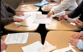 Students seated at grouped desks with folded paper products, folding instruction handouts, and cost worksheets spread in front of them.