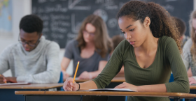 Students working independently at their desks, writing at an assessment in a classroom with a chalkboard in the background.