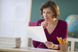 Teacher seated at a desk holding a printed page of data, pencil in hand, reviewing it closely.