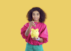 A smiling teenage girl with curly hair, wearing a bright pink top and green pants, drops a coin into a yellow piggy bank that she holds in her open palm against a solid yellow background.