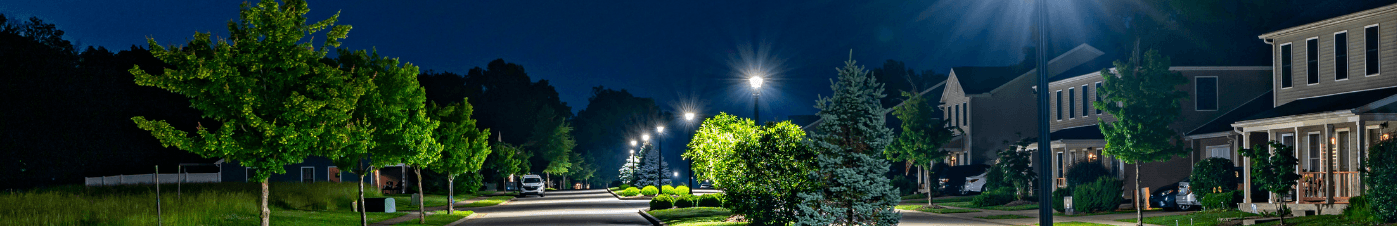 A residential street at night illuminated by a row of bright streetlights, with trees and houses visible on either side under a deep blue sky.