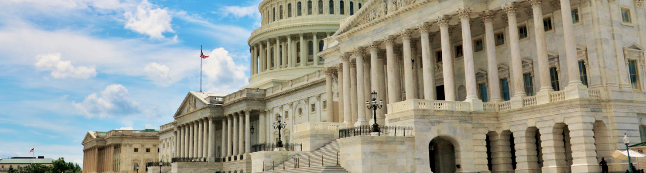 The U.S. Capitol building on a sunny day, with its iconic dome, white marble columns, and steps visible under a partly cloudy blue sky.