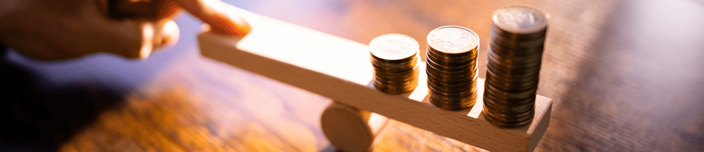 A hand tips a wooden balance beam holding three stacks of coins of increasing height, representing the balancing act of the inflation rate and federal funds rate explored in this lesson.