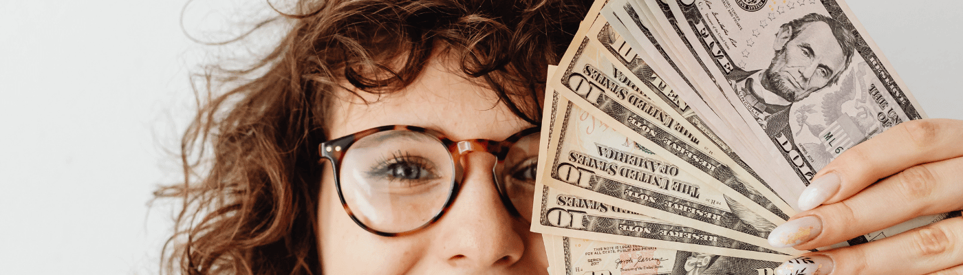 A young woman with curly hair and glasses peers over a fan of U.S. dollar bills, representing the personal financial decision-making skills developed through A Practical Guide to the U.S. Economy.