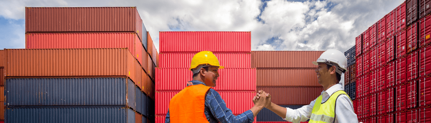 Two workers in hard hats shake hands in front of stacked shipping containers, representing the trade agreements and worker support programs explored in this lesson.