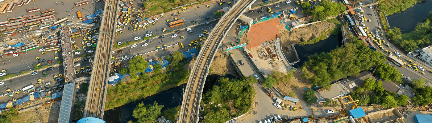 9:08 AMAlt text: An aerial view of intersecting highways and rail lines under construction amid dense urban traffic, representing the infrastructure investments that drive economic growth explored in this lesson.