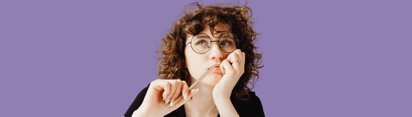 A young woman with curly hair and glasses holds a pencil thoughtfully against her lips while looking upward, against a purple background, representing the economic thinking and proposal-writing students engage in during this assessment lesson.
