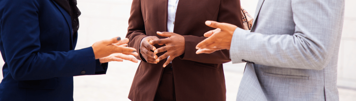 Three professionals in business attire standing in a group discussion with hands gesturing expressively, representing the presentation and debate of policy proposals and economic recommendations.
