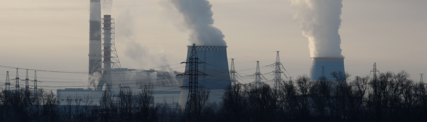 A coal power plant with multiple smokestacks and cooling towers releasing large plumes of smoke and steam into a hazy sky, with power line towers and bare trees silhouetted in the foreground.
