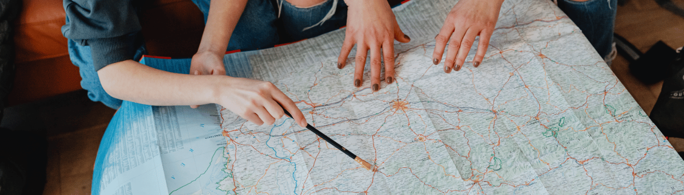 Two people leaning over a large road map spread across a table, with one pointing to a location using a pencil, representing the process of navigating and analyzing economic data.