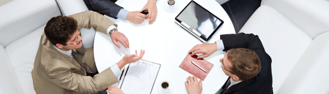 An overhead view of three people in business attire seated around a round table, debating documents and a tablet, representing the analysis of multiple perspectives on economic policy.
