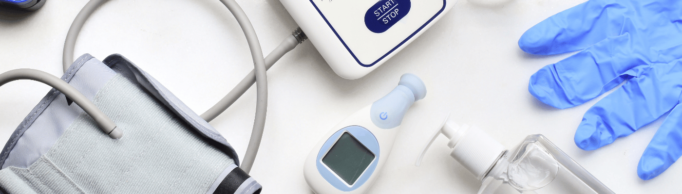 A blood pressure monitor, digital thermometer, pulse oximeter, hand sanitizer, and blue latex gloves arranged on a white surface — tools for measuring and monitoring health.
