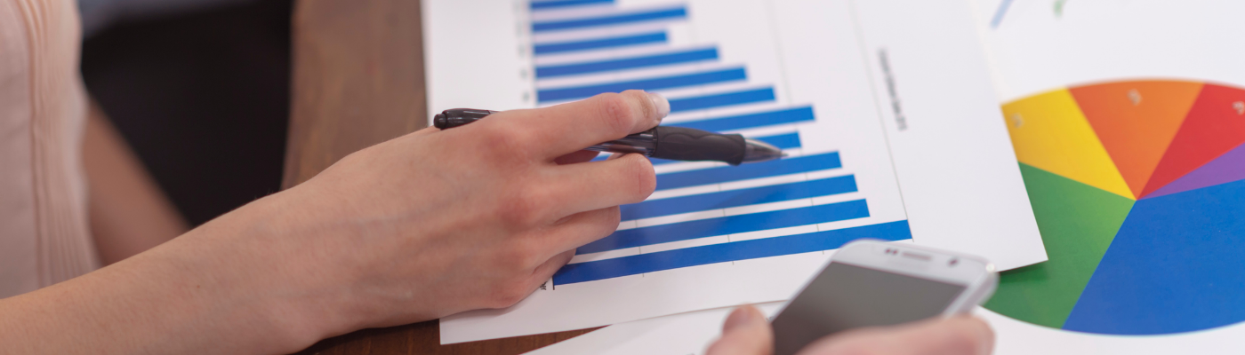 A person holding a pen and pointing to a printed bar chart, with a colorful pie chart visible alongside it, representing the analysis of economic data.